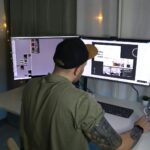 man in green dress shirt and black cap sitting in front of computer